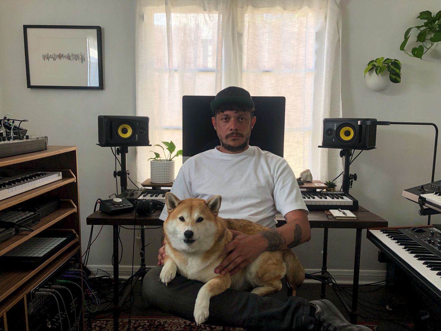 Alain Emile, Pablo Swiss, seated in his studio with a Shiba Inu on his lap, keyboards and monitors around him, a curtained window behind