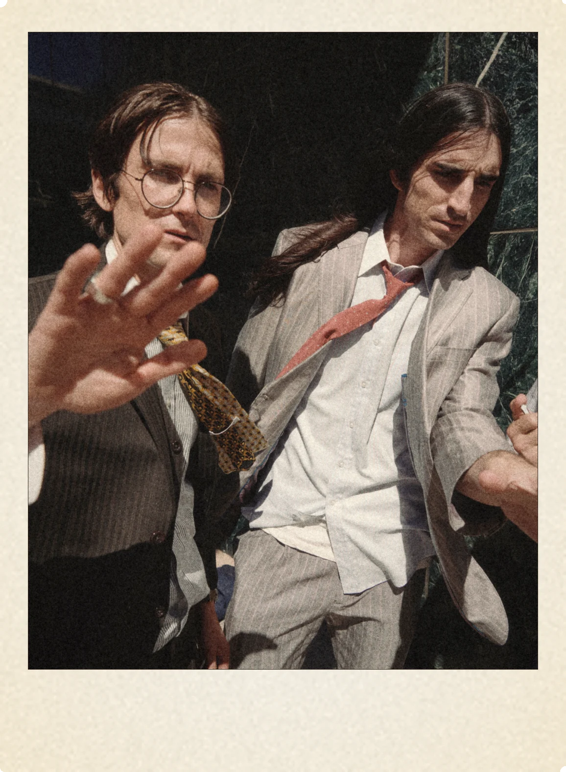 Zachary Cole Smith and Colin Caulfield of DIIV in suits against dark marble, a hand reaching toward the lens; photograph by Louie Kovatch, 2026